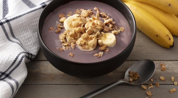 Fotografia em tons de branco e preto de uma bancada branca, sobre ela um recipiente redondo preto com açaí, um cacho de banan