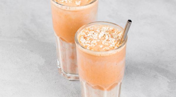 Fotografia de dois copos de vidro com um suco de pitanga com melaço de cana. Dentro dos copos com o suco, um canudo de metal, e embaixo dos copos, uma mesa cinza em tom claro.