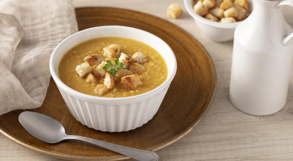 Fotografia em tons de bege de uma bancada, um prato marrom com um potinho branco com a sopa e pequenas torradinhas. Ao lado uma colher prata e do outro lado um pote com pedacinhos de torrada e uma jarra branca.
