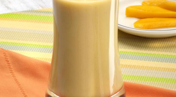 Fotografia em tons de laranja em uma bancada de madeira com uma toalha colorida listrada, um pano laranja e um copo de vidro alto com a vitamina de banana com cupuaçu. Ao fundo, um pratinho branco raso com fatias de manga.