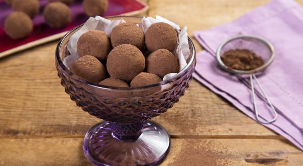 fotografia em tons de marrom e lilás de uma bancada marrom vista de cima, contém uma taça lilás com trufas redondas de chocolate e ao lado um pano lilás com uma peneira que contém cacau em pó.