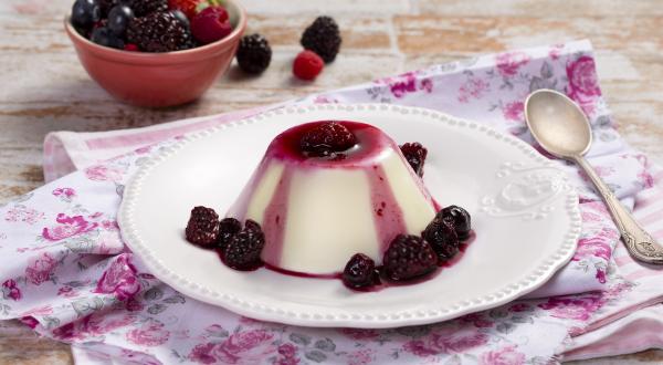 fotografia em tons de branco e roxo, contém um pano branco com rosas desenhadas, acima um prato redondo branco com um mini Flan com calda de frutas vermelhas e frutas vermelhas em volta.