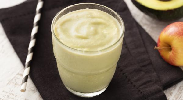 Fotografia em tons de branco e preto de uma bancada de madeira branca com um paninho preto, sobre ele um copo de vidro com o creme de abacate e um canudo. Ao fundo meio mamão e uma maçã.