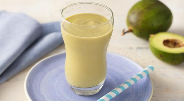 Fotografia em tons de azul e verde de uma bancada com um pratinho azul, um copo transparente com uma vitamina de abacate e um canudo. Ao fundo um paninho azul e um abacate.