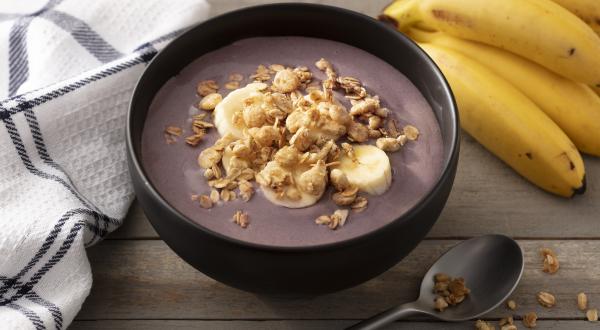 Fotografia em tons de branco e preto de uma bancada branca, sobre ela um recipiente redondo preto com açaí, um cacho de banana, uma colher e um paninho branco com listras pretas.