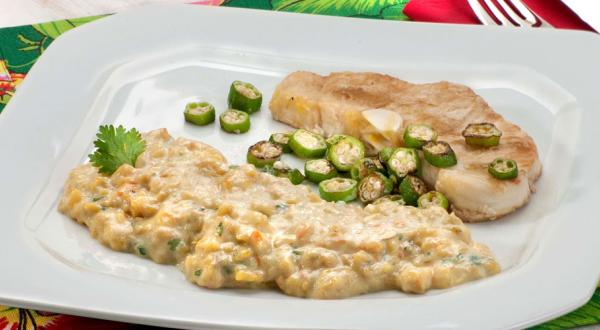 Fotografia em tons de verde em uma mesa com uma toalha verde florida, um prato branco grande com o peixe servido com quiabo e com purê de banana da terra.