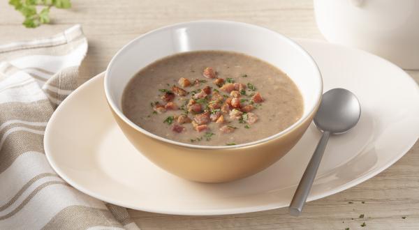Fotografia em tons de bege de uma bancada com um prato oval, sobre ele um bowl bege com a sopa de feijão e pedacinhos de bacon. Ao lado uma colher prateada. Ao redor um pano listrado branco e bege, ramo de salsinha e um pote branco.