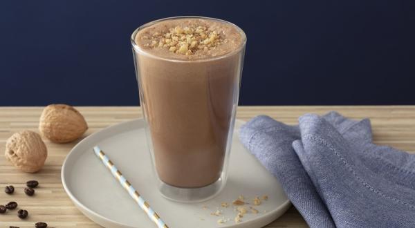 Foto em tons de marrom da receita de smoothie de café com nozes servida em um copo alto sobre uma base de porcelana cinza com um canudo listrado ao lado. Como decoração há duas nozes e um paninho azul