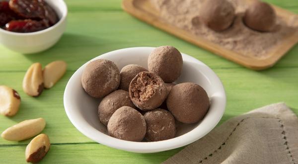 Fotografia em tons de marrom em uma bancada de madeira de cor verde. Ao centro, um bowl branco contendo as trufas. Ao lado, há algumas castanhas do pará e um pano marrom. Ao fundo, um pote com tâmaras e uma tábua com mais trufas.