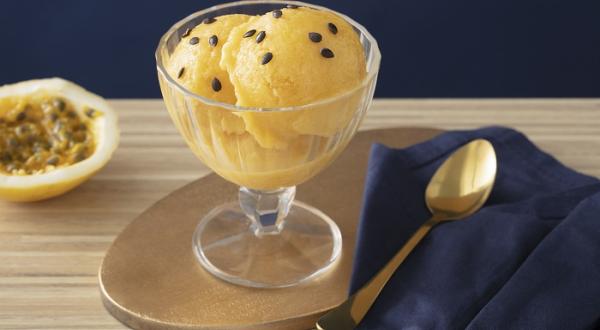 Foto em tons de amarelo da receita de sorbet de damasco e maracujá servida em uma taça de vidro sobre uma mesa de madeira com uma colher prateada ao lado sobre um pano de veludo azul. Ao fundo ainda há metade de um maracujá