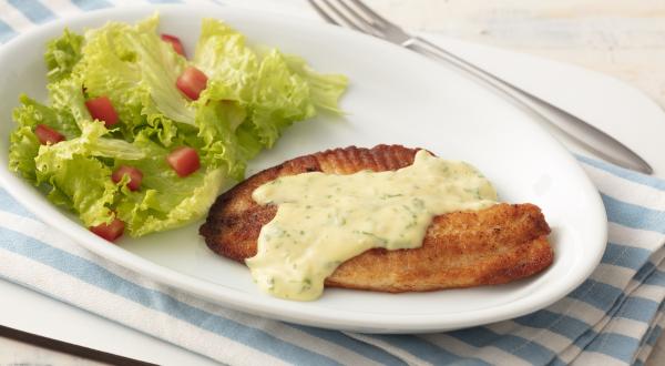 Fotografia em tons de azul em uma bancada de madeira clara com um pano listrado azul claro e um prato oval branco com o filé de pescada com molho de limão e mostrada e salada de alface com tomates.