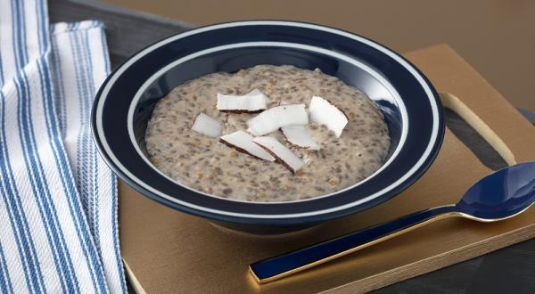 Foto em tons de marrom da receita de mingau de sementes e coco servida em um prato fundo preto com uma colher prateada ao lado sobre uma tábua de madeira lisa com um paninho azul ao lado