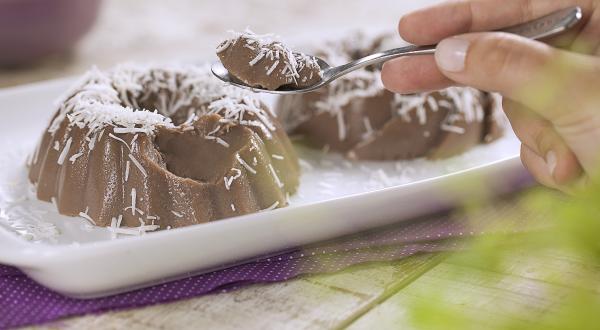 .Fotografia em tons de roxo e branco de uma bancada de madeira branca com um paninho roxo, sobre ele um prato retangular com dois gelados de dark chocolate. Sobre ele uma colher com um pedaço do gelado. Ao fundo um recipiente redondo com coco.