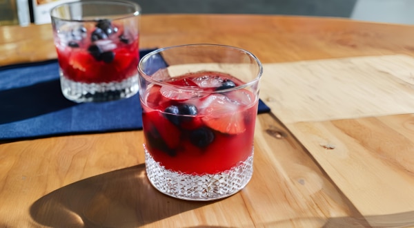 Fotografia de dois copos de vidro baixos com uma bebida sem álcool com frutas vermelhas e gelo. Os copos estão afastados um do outro, e o copo do fundo está sobre um pano azul de tom escuro, sobre uma mesa de madeira.