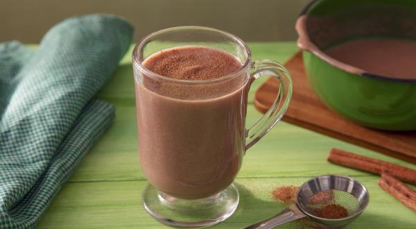 Fotografia em tons de marrom em uma bancada de madeira de cor verde. Ao centro, uma xícara contendo o chocolate quente. Ao lado, há uma toalha verde quadriculada e ao fundo, uma tábua de madeira com uma panela verde em cima.