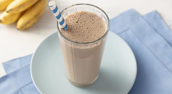 Fotografia em tons de azul e marrom de uma bancada com um paninho azul dobrado, um prato azul e um copo com a bebida de chocolate e um canudo. Ao fundo um cacho de bananas.