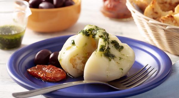 Fotografia em tons de branco em uma bancada de madeira de cor branca. Ao centro, um prato azul contendo a burrata com um garfo ao lado. Ao fundo, está uma cesta com pães e tomates secos.