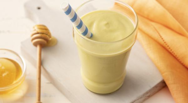 Fotografia em tons de amarelo e verde em uma bancada de madeira clara com um pano laranja, ao centro uma tábua de madeira com um copo de vidro com a vitamina de abacate dentro e um canudo de papel com listras azuis. Ao lado, um potinho de mel.