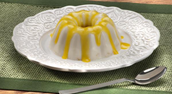 Fotografia em tons de amarelo e verde em uma bancada de madeira com uma toalha verde, um prato redondo branco raso e o flan de coco com calda de pequi em cima dele. Ao lado, uma colher.