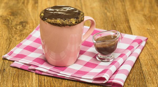 Fotografia em tons de rosa em uma bancada de madeira escura, um pano rosa xadrez, uma caneca rosa com um bolo de caneca de banana com mel, aveia e cobertura de chocolate. Ao lado, um potinho de vidro com a calda de chocolate.