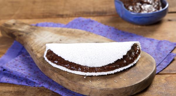 Fotografia em tons de marrom e roxo de uma bancada de madeira com um paninho roxo, sobre ele um tábua de madeira com uma tapioca. Ao fundo um recipiente roxo com creme de dark chocolate.