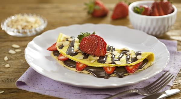 Fotografia em tons de lilás em uma bancada de madeira, com um paninho lilás com bolinhas, um prato branco raso com a crepioca recheada de chocolate e morangos.