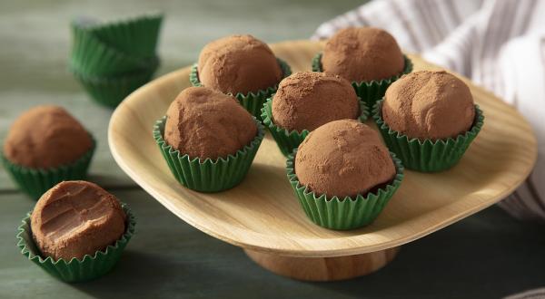 Fotografia em tons de marrom em uma bancada de madeira de cor verde. Ao centro, um prato de madeira contendo os brigadeiros. Ao, há um brigadeiro mordido e fundo, um pano branco.