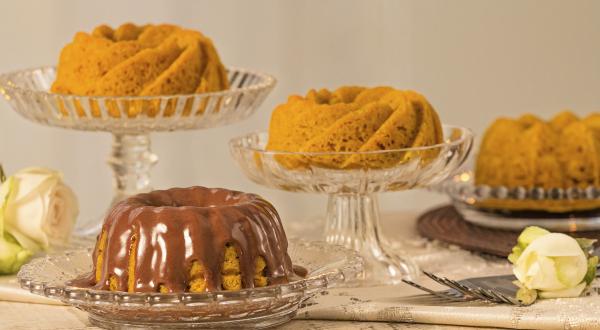 fotografia em tons de cinza, laranja e marrom vista de frente. Contém 4 bolos de cenoura e um deles esta com calda de chocolate, todos estão dentro de recipientes de vidros