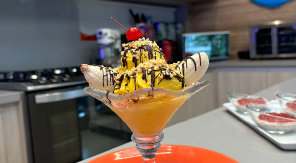 Fotografia de uma taça de vidro com banana split com calda de chocolate, farofa de castanha e sorvete de abacate.