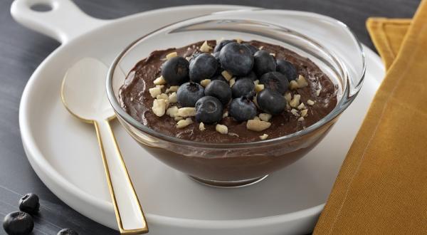 Foto em tons escuros da receita de mousse de chocolate com mirtilo servida em uma taça de vidro sobre uma frigideira de porcelana branca com uma colher branca ao lado