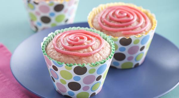 Fotografia em tons de azul e rosa em uma mesa de madeira azul, um pano rosa, um prato azul redondo com dois cupcakes de morango com aveia em forminhas coloridas.