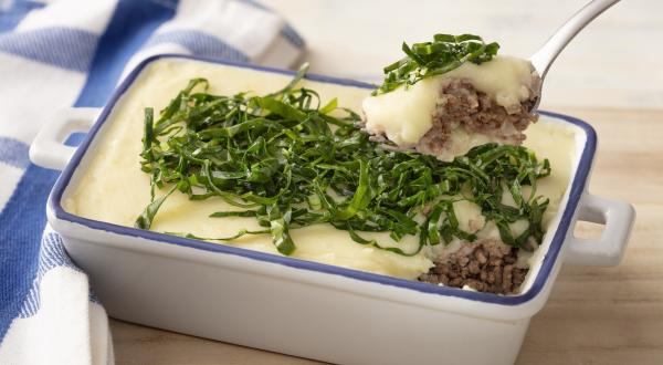 Fotografia em tons de azul em uma bancada e madeira, um pano xadrez azul e ao centro um recipiente refratário branco com detalhes em azul e o escondidinho de carne e batata e couve por cima para decorar.