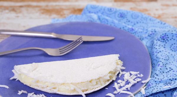 Fotografia em tons de azul em uma bancada de madeira clara, um pano azul, um prato azul redondo com uma tapioca de coco em cima dele.