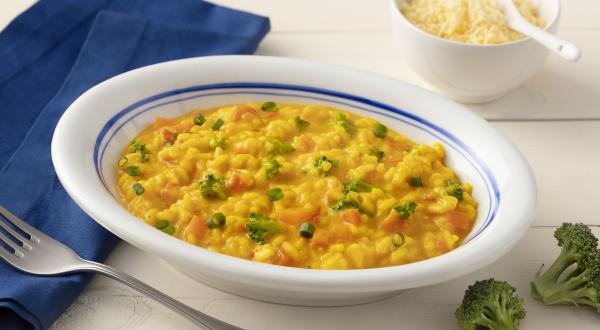 Fotografia em tons de azul em uma bancada de madeira clara com um pano azul escuro ao lado e um prato branco com detalhes em azul ao centro com o risoto de legumes com cúrcuma dentro. Ao lado, um potinho branco com queijo parmesão ralado e dois brócolis.