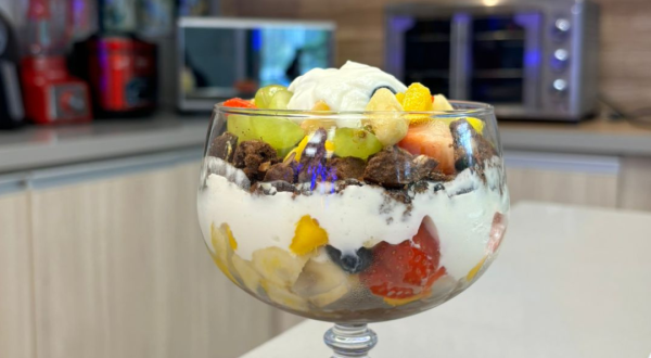 Fotografia de uma taça grande de vidro com pavê de frutas e iogurte, O pavê está sobre uma bancada e no fundo tem aparelhos eletrodomésticos.