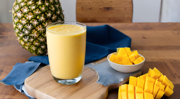 Fotografia de um copo de vidro com smoothie de iogurte, manga e abacaxi. O copo está sobre uma mini tábua de madeira, ao lado de dois recipientes brancos pequenos com pedaços de manga. No fundo tem um abacaxi inteiro sobre um pano azul escuro.