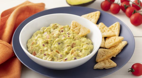 Fotografia em tons de azul em uma bancada de madeira clara com um pano laranja ao lado e um prato grande azul e um prato fundo branco com o guacamole dentro. Ao lado, um pedaço de avocado, alguns tomatinhos e biscoitinhos.