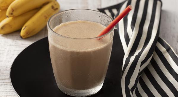 Fotografia em tons de branco e preto de uma bancada de madeira branca com um prato preto, sobre ele um copo de vidro com a bebida cremosa e um canudo vermelho. Ao lado um paninho listrado branco e preto e um cacho de banana.