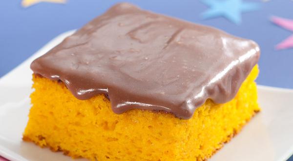 Fotografia em tons de azul, laranja e marrom de uma bancada azul, sobre ela um prato branco quadrado com uma fatia de bolo de cenoura com cobertura de chocolate. Ao lado um garfo verde. Ao fundo uma estrela azul e uma rosa.