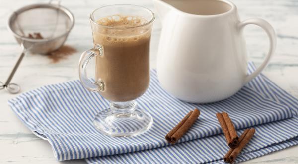 Fotografia em tons de azul em uma bancada de madeira clara com um pano azul com listras finas e ao centro uma taça com o cappuccino e ao lado uma jarrinha branca. Ao lado, três paus de canela e um difusor com o chocolate em pó.