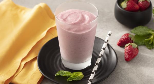 Fotografia em tons de amarelo, branco e preto de uma bancada branca com um prato preto redondo, sobre ele um copo de vidro com smoothie de morango e um canudo branco com bolinhas pretas. Ao lado um paninho laranja e morangos.