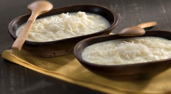 Fotografia em tons de marrom, branco e bege, com dois recipientes de madeira escura com tapioca, coco ralado e duas colheres de pau sobre guardanapo bege, tudo sobre bancada em tons de cinza.