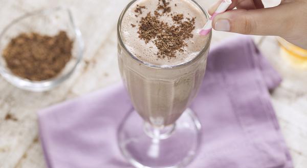 Fotografia em tons e branco e roxo de uma bancada branca com um paninho roxo, sobre ele uma taça de vidro com milk shake e um canudo. Ao lado um recipiente de vidro com raspas de chocolate.