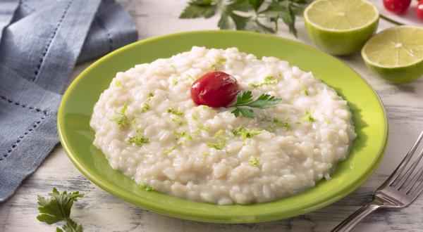 Fotografia em tons de verde em uma bancada de madeira de cor branca. Ao centro, um prato verde contendo o arroz. Ao lado, há um garfo e um pano azul. Ao fundo, há um limão partido ao meio, tomate cereja e salsinha espalhados.