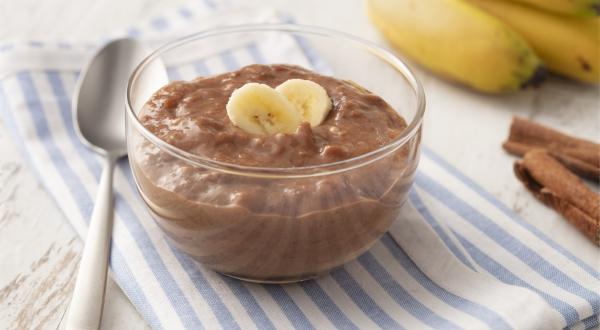 Fotografia em tons de azul em uma bancada de madeira clara com um paninho listrado azul e em cima um potinho de vidro com o doce de banana e cacau. Ao lado, algumas bananas, uma colher e canela em pau.