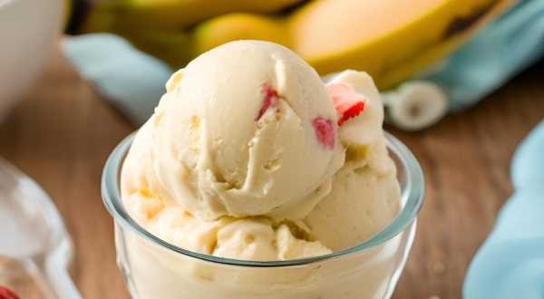 Foto da receita de Sorvete de Banana com Morango. Observa-se uma tacinha de vidro com as bolas de sorvete dentro, na cor amarela e com pedacinhos de morango. Foto gerada por IA.