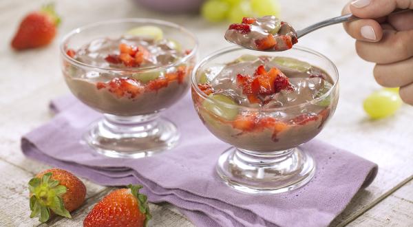 Fotografia em tons de branco e roxo de uma bancada de madeira branca com um paninho roxo, sobre ele duas taças de frutas com creme e uma colher retirando um pedaço. Ao fundo uvas e morangos. A frente dois morangos.