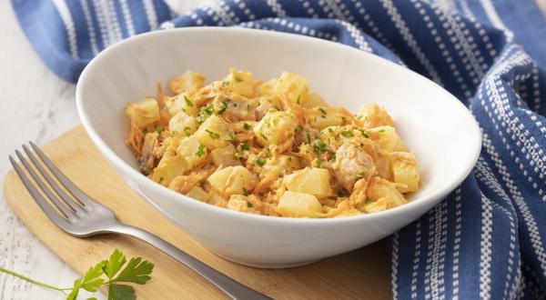 Fotografia em tons de azul em uma bancada de madeira com um pano listrado azul ao lado. No centro, um prato fundo branco com a salada de batata, atum, cebola e salsinha.