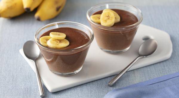 Fotografia em tons de azul e marrom de uma bancada com um paninho azul, duas tacinhas transparentes com o creme de chocolate e rodelas de banana decorando, ao lado duas colheres pratas e ao fundo 3 bananas.