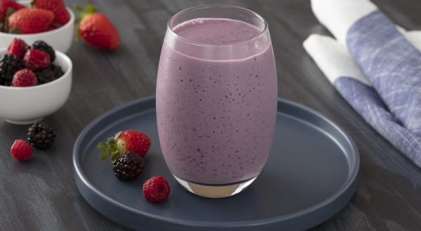 Foto em tons de roxo da receita de shake de frutas vermelhas com chia servida em um corpo largo sobre uma base de porcelana azul escura circular com frutas vermelhas ao lado. Ao fundo, há dois recipientes com frutas vermelhas e um pano azul.